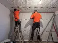 Dwóch mężczyzn w pomarańczowych koszulkach montuje materiał izolacyjny, pokazując, jak wygłuszyć ścianę.