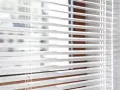 Close-up of white horizontal blinds with visible cords, showing the slats and a hint of the window frame.