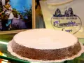 A Torta Caprese dusted with powdered sugar sits on a glass counter, with a painting and shop logo in the background.