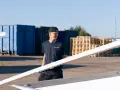 Man i mörkblå t-shirt med "ISOLERAB" logga bär en träskiva. Bakom honom syns blå containrar och träpallar.