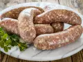 Kilka surowych kiełbasek białych parzonych ułożonych na marmurowej desce, ozdobione gałązką zielonych liści.