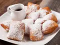 Une assiette de beignets saupoudrés de sucre glace, parfaits pour une recette pâte à beignets salés. Un petit pichet de sauce est à côté.