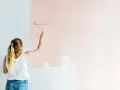 Frau streicht mit einer Malerrolle eine Wand in einem zarten Rosaton. Die Wohnung bekommt einen neuen Anstrich.