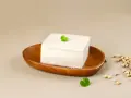 Kostka białego tofu na drewnianej miseczce, ozdobiona listkiem. Obok garść ziaren soi. To właśnie co to tofu – produkt z soi.