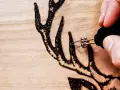 Dettaglio di un disegno a pirografia su legno: una mano guida le punte pirografo per creare le corna di un cervo.