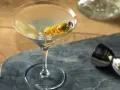 Elegancki martini z wódką, ozdobiony trzema oliwkami na wykałaczce, gotowy do degustacji. Obok metalowy jigger.