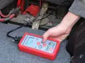 Ręka trzyma tester akumulatora DBT-12+, pokazujący 12.14V. To prosty sposób, jak sprawdzić akumulator w samochodzie.
