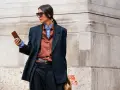 Mujer con estilo combinando traje negro con camisa marrón y azul, gafas de sol y móvil en mano.
