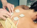 Fizjoterapeuta umieszcza elektrody na plecach pacjentki, przygotowując do zabiegu TENS. Należy pamiętać o przeciwwskazaniach do jego stosowania.