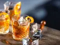 Tre old fashioned-drinkar med is och apelsinskal på ett träbord. En cocktailgaffel syns i bakgrunden.