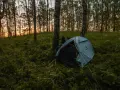 Namiot nad jeziorem, otoczony drzewami i trawą, czeka na noc. Zachód słońca maluje niebo na pomarańczowo.