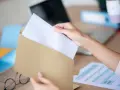 Dłoń wkłada biały papier do brązowej koperty. Wzór adresowania koperty A4 jest gotowy do wypełnienia.