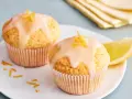 Zwei einfache Zitronenmuffins mit Zuckerguss und Zitronenschale garniert, daneben ein Zitronenschnitz.