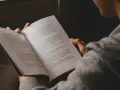 Osoba czyta książkę, skupiona na treści. W jej dłoniach spoczywa otwarta księga, a jej wzrok skierowany jest na strony, gdzie słowa tworzą historię, być może o trudnych wyborach i ich konsekwencjach, gdzie **inwersja** losu jest na porządku dziennym.