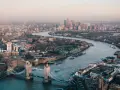 Stunning drone photography and videography captures London's iconic Tower Bridge and the winding Thames River, with the city skyline stretching into the hazy distance.
