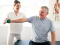 Mann hebt Hantel unter Anleitung einer Therapeutin. Physiotherapie nach Bobath hilft bei der Rehabilitation.
