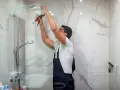 Instalator w granatowym ogrodniczkach dokręca słuchawkę prysznicową. Biały montaż w nowoczesnej łazience.
