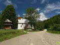 Drewniana łopienka i biały kościółek w otoczeniu zieleni. Droga prowadzi do lasu, a na niej dwie postacie.