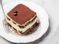 Una fetta di tiramisù, preparata con 500 g di mascarpone, guarnita con cacao e chicchi di caffè, su un piatto bianco con forchetta.