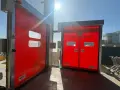Due porte rosse automatiche, una aperta, mostrano come alzare una porta dal pavimento. Luce solare intensa.