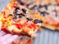 Kawałek pysznej pizzy funghi z pieczarkami, cebulą i pomidorami, trzymany w dłoni.
