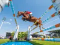 Un magnifique cheval de course brun saute par-dessus un obstacle coloré, sous un ciel bleu clair, lors d'une compétition équestre devant une foule attentive.