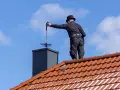 Kominiarz w czapce na dachu czyści komin. Zastanawiasz się, ile kosztuje przegląd kominiarski?