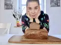 Kobieta z dłońmi podpartymi pod brodą patrzy na pyszny chleb wieloziarnisty. Jeden bochenek leży na drugim, na drewnianej desce.