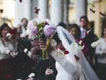 Płatki róż sypią się na młodą parę. W tle widać gości, a na pierwszym planie bukiet panny młodej. To moment, gdy ksiądz na ślubie błogosławi nowożeńców.