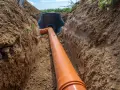 Pomarańczowa rura kanalizacyjna prowadzi do czarnego zbiornika. Trwa podłączenie do istniejącego przyłącza w wykopie.