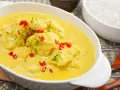Delizioso pollo al curry con latte di cocco, guarnito con peperoncino rosso e erba cipollina. Accanto, una ciotola di riso bianco.