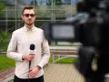Młody dziennikarz w okularach trzyma mikrofon z logo "FLESZ" przed kamerą.