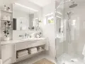 Une petite salle de bain moderne et lumineuse avec une douche vitrée, un lavabo suspendu et des étagères remplies de plantes et de produits de soin.