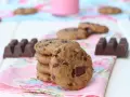 Una pila de galletas de chocolate caseras, con trozos de chocolate visibles, junto a barras de chocolate y un vaso de leche rosa.