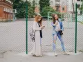 Dwie dziewczyny w modnych stylizacjach, idealnych na outfit do szkoły, stoją przy płocie. Jedna robi sobie selfie, druga przegląda telefon.