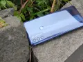 Błyszczący tył telefonu **Moto G 50** z potrójnym aparatem, leżący na kamieniach wśród zieleni.