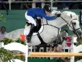 Une cavalière en tenue bleue et blanche saute un obstacle avec son cheval gris. Le cheval, nommé Levisto Z, montre une puissance impressionnante.