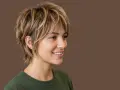 Donna con capelli scalati biondi, un taglio moderno che anticipa i tagli capelli 2025 medi. Sorriso radioso su sfondo marrone.