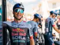 Cyclist smiles on a vuelta rest day, wearing a BORA-hansgrohe kit with Red Bull logos. Other team members and bikes are visible.