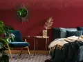 Habitación con pared de pintura a la tiza roja, sillón azul, cama con mantas y cojines, espejo sol y planta.