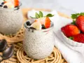 Zwei Gläser mit cremigem Chia Pudding, garniert mit Erdbeeren, Bananen und Blaubeeren. Daneben eine Schale frischer Erdbeeren.