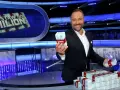 Uśmiechnięty prowadzący teleturniej trzyma w dłoni paczkę pieniędzy. Na stole piętrzą się stosy banknotów.