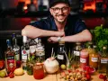 Uśmiechnięty mężczyzna w czapce i okularach za ladą pełną alkoholi i owoców. To przyszłość: drink bar zamiast barmana.