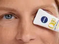 Kobieta z niebieskimi oczami, z kremem Nivea Q10 Anti-Wrinkle Power zakrywającym jedno oko. Skóra wokół oczu ma drobne kurze łapki.