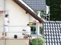 Dwóch mężczyzn na rusztowaniu maluje białą elewację domu. Sprawdź, jaka jest malowanie elewacji cena.