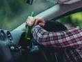 Mężczyzna w kratę trzyma butelkę, opierając głowę na kierownicy. To obraz ostrzegawczy przed jazdą pod wpływem alkoholu, przypominający o zagrożeniach i potrzebie blokady alkoholowej.