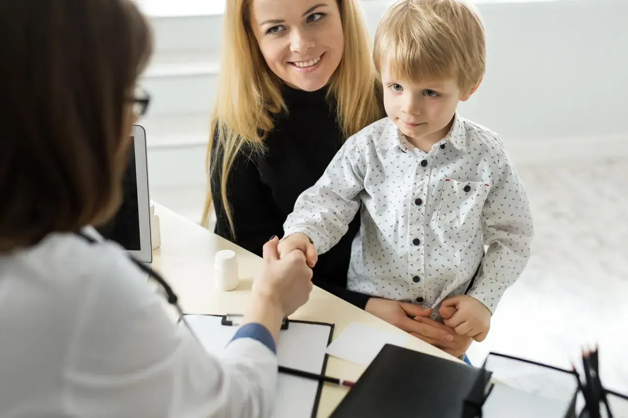 Mama z synkiem u lekarza. Chłopiec ściska dłoń lekarce, a mama uśmiecha się. W tle widać tablet i białą buteleczkę, jakby z lekiem. To ważny moment, być może wpis do książeczki zdrowia.