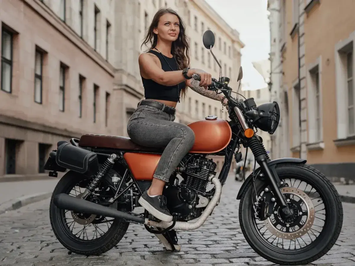 Kobieta w czarnym topie i jeansach siedzi na pomarańczowym motocyklu na brukowanej ulicy. To idealna kategoria a dla miłośniczek wolności.