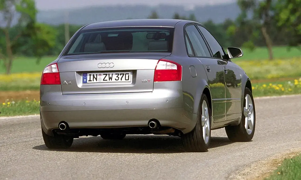 Srebrne Audi B6 sedan z tablicą rejestracyjną IN ZW 370 jedzie po asfaltowej drodze wśród zieleni i żółtych kwiatów.