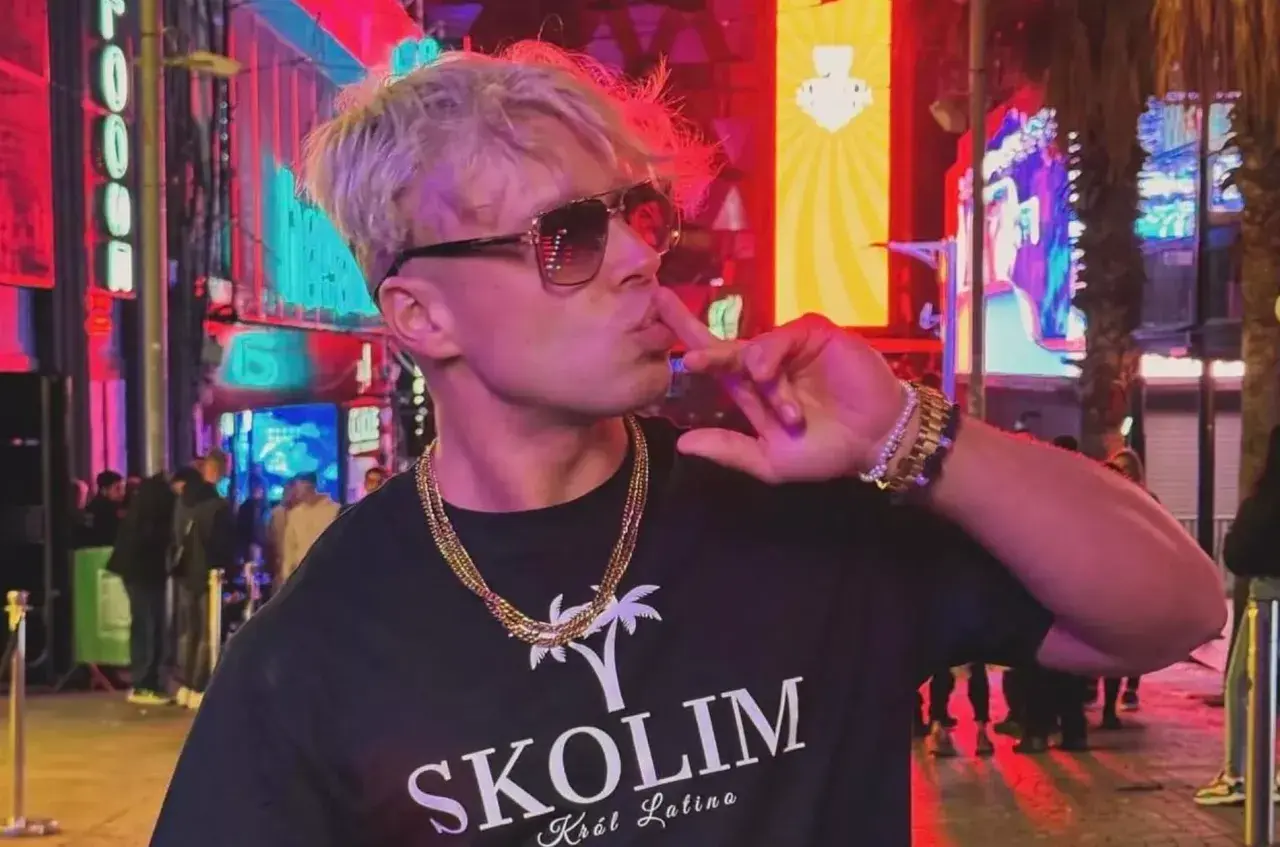 Skolim w okularach i złotych łańcuchach, pozuje na tle neonów. Król Latino w swoim wieku, w blasku miasta.